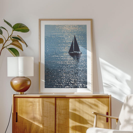 Framed sailboat art print displayed in a bright, modern interior with mid-century styling. The artwork features a dark sailboat on shimmering blue water, with sparkling light reflections in an abstract dot pattern. Set above a light wood credenza beside a ceramic table lamp and leafy plant, the print adds a serene, coastal feel to the sunlit room.