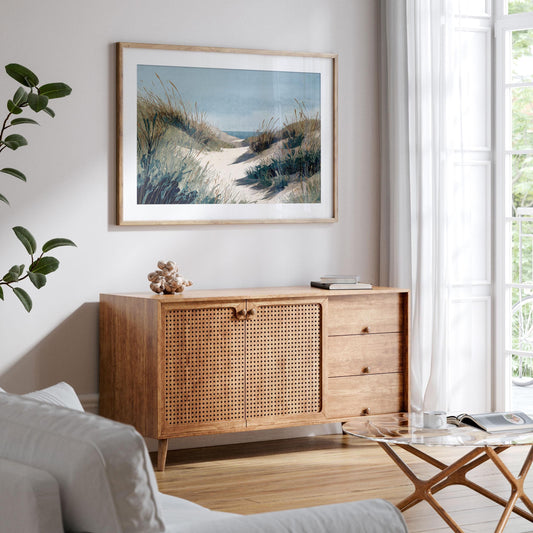 Framed coastal watercolor print of a sandy path through grassy dunes toward the ocean, displayed above a wooden cabinet with woven doors in a bright, airy living room. Decor includes a glass coffee table, white sofa, potted plant, and sheer curtains letting in natural light.