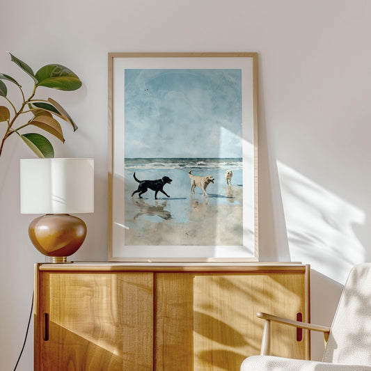 Framed watercolor-style print of three Labrador retrievers playing on a beach, hung above a light wood cabinet in a sunlit room. A black lab approaches two yellow labs, one holding a stick, with reflections in wet sand under a blue sky and ocean waves. A ceramic lamp and leafy plant sit on the cabinet, with sunlight casting soft shadows on the wall.