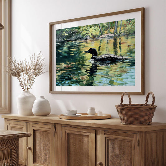 Framed print of a loon swimming on a reflective lake, surrounded by lush green foliage and warm sunlight. The painting’s rich greens, golds, and blues bring a sense of calm and nature indoors. Displayed above a rustic wooden sideboard with neutral ceramics, wicker basket, and dried grasses, the scene creates a cozy, natural aesthetic in a softly sunlit room.