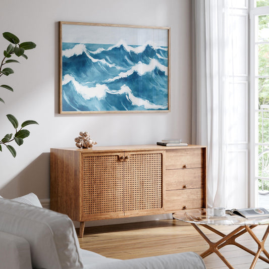 Framed ocean wave painting displayed above a mid-century modern sideboard in a bright, coastal-inspired living room. The artwork features layered blue waves with foamy white crests, adding a calming, nautical touch to the neutral space with wood and rattan textures.