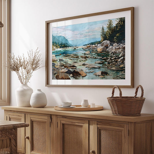 Framed watercolor landscape of a rocky river and pine forest hangs above a natural wood sideboard in a softly lit room. The painting shows turquoise water flowing past stones toward misty blue mountains under a pink-tinged sky. The decor includes white ceramic vases, a wooden tray with a candle and cup, and a woven basket, creating a warm, minimalist, nature-inspired aesthetic.