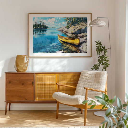 Framed watercolor-style print of a yellow canoe on a bright blue lake hangs above a mid-century modern wood sideboard. A sculptural tan vase, cozy cream armchair, tall floor lamp, and leafy plant complete the sunlit living room. The artwork shows rocky shore, pine trees, and clear sky reflected in the calm water.