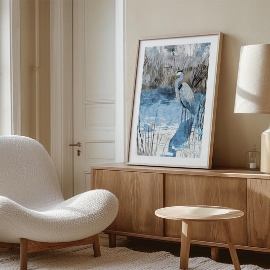 Large framed art print of a heron standing in blue water with reeds, displayed on a modern wooden sideboard in a cozy living room. The artwork features cool blue, gray, and earthy tones, adding a calming nature-inspired accent to the neutral décor with soft lighting, a white textured lounge chair, and a minimalist lamp.