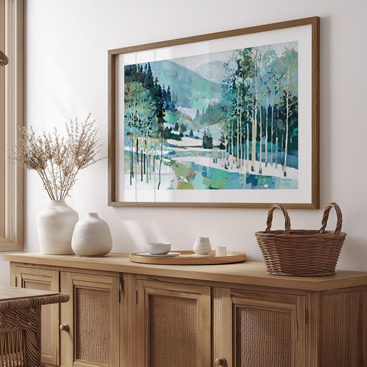 Framed painting of a snowy mountain forest scene in cool tones of blue, green, and white, hanging on a wall above a rustic wooden sideboard. The artwork features tall birch trees, layered hills, and a geometric patchwork base. Decor includes white ceramic vases with dried stems, a wooden tray with cups, and a woven basket, creating a cozy, natural home setting.