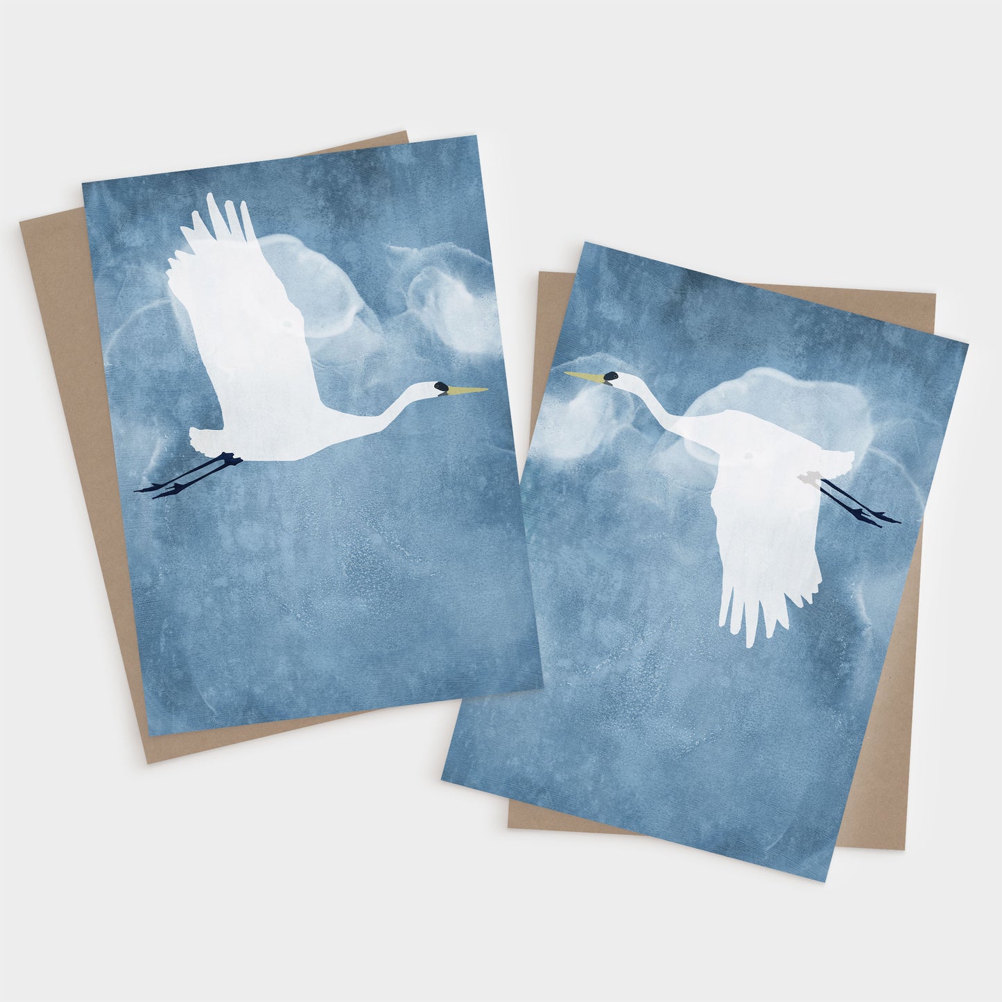 Set of two greeting cards featuring minimalist illustrations of white egrets in flight against a textured blue sky, one with wings raised and the other with wings down, each paired with a kraft brown envelope.
