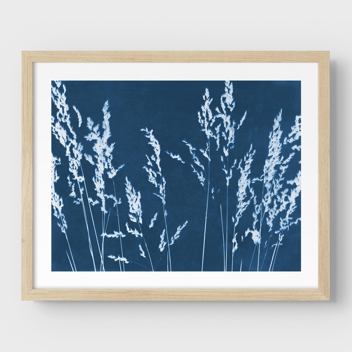 Framed cyanotype print featuring white wild grass silhouettes against a deep blue background, presented with a natural wood frame and white border.