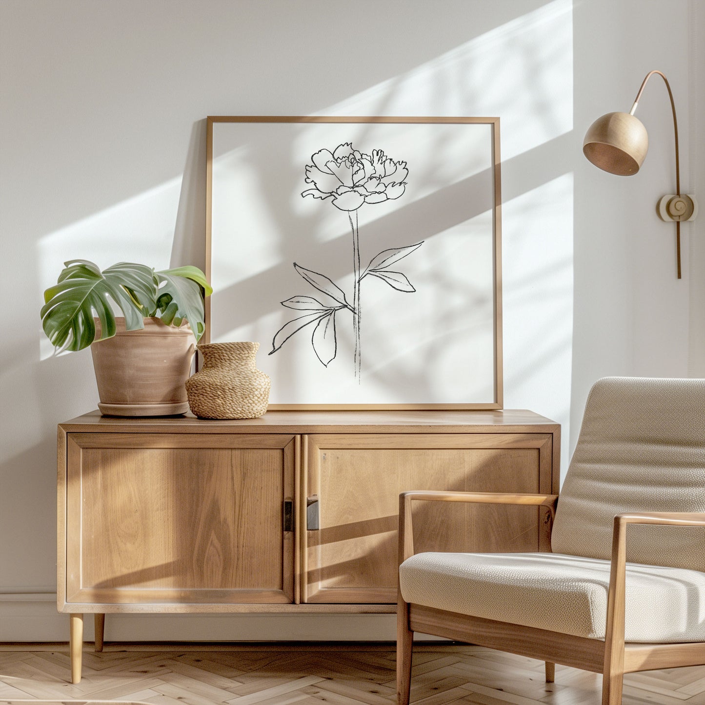 Framed line drawing of a peony flower on a wooden sideboard, styled with a potted monstera, woven vase, and a beige armchair in a sunlit modern interior.