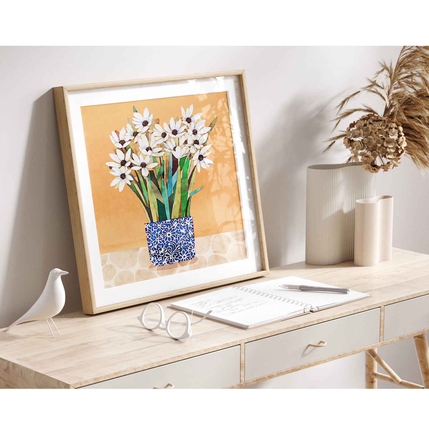 A framed print of white flowers in a blue patterned vase against an orange background, styled on a light wood desk with decorative vases, a bird figurine, and a notebook, illuminated by soft natural light.