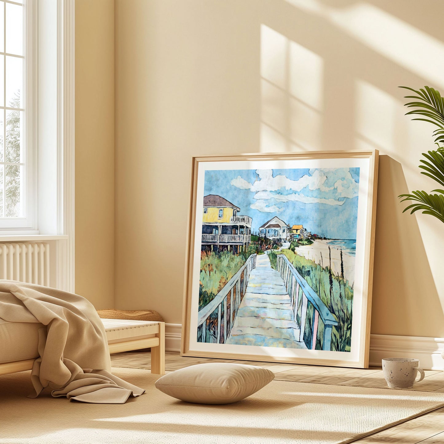 Framed beach house painting leaning against a sunlit wall in a cozy, minimalist room. The artwork shows a boardwalk leading through grassy dunes toward colorful beachfront houses, including a prominent yellow stilt house, with the ocean and blue sky in the background.