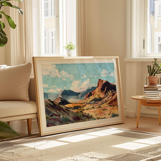 Large framed painting of a desert canyon with red rock cliffs, rolling hills, and distant blue mountains under a bright sky with scattered clouds, resting on the floor in a sunlit living room beside a cream sofa, potted plants, and a small table stacked with books.