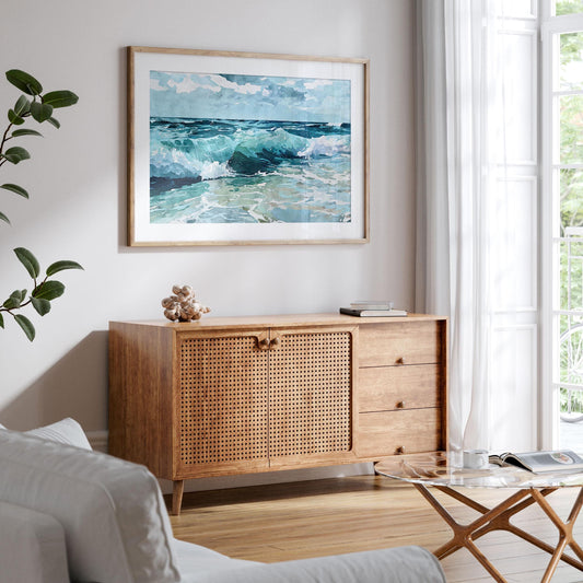 Framed watercolor painting of turquoise ocean waves hangs above a mid-century modern wooden cabinet in a bright living room with light wood floors, white walls, a glass coffee table, and large windows with sheer curtains.