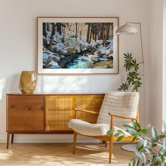 Framed winter forest painting displayed in a bright, modern living room. The artwork depicts snow-covered rocks, tall trees, and a reflective stream in cool blues and warm golden tones. It hangs above a mid-century wooden sideboard with a geometric vase, beside a cozy cream armchair and leafy plant, creating a serene, stylish atmosphere.