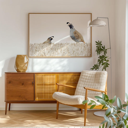 Framed watercolor print of two California quail in tall beige grass hangs above a mid-century modern wood sideboard with a geometric vase, next to a cozy cream armchair and floor lamp in a bright, minimalist living room.