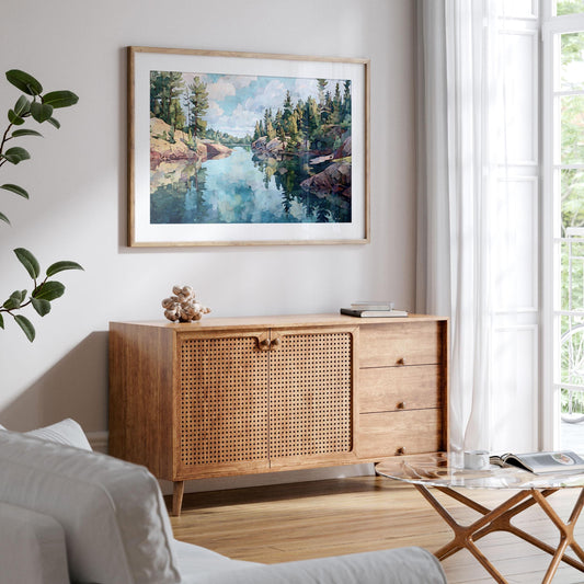 Framed landscape painting of a northern Minnesota lake hangs above a mid-century style wood sideboard with woven rattan doors. The artwork shows calm blue-green water reflecting pine trees and rocky shores under a cloudy sky. Room features white walls, a light sofa, glass coffee table, and a large window with sheer curtains, creating a bright, airy, modern space.