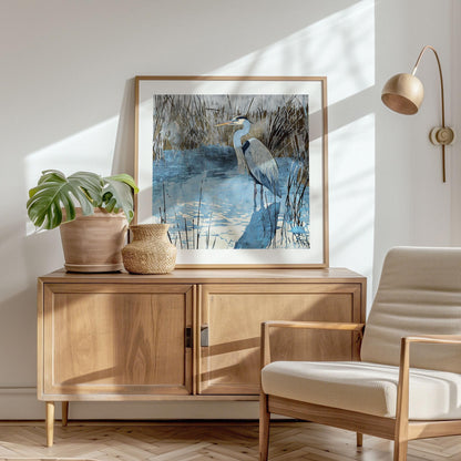 Framed heron art print with blue and gray tones, showing the bird standing in reflective water among reeds. Displayed on a wooden sideboard in a sunlit modern living room, the scene is accented with a potted monstera, woven vase, cream armchair, and a minimalist wall lamp, creating a calm, natural atmosphere.