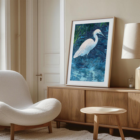 Framed watercolor art print of a white egret wading in rippling blue water with green reeds, displayed leaning against a wall on a wooden sideboard in a modern living room with a textured white chair, round wood table, and neutral decor.
