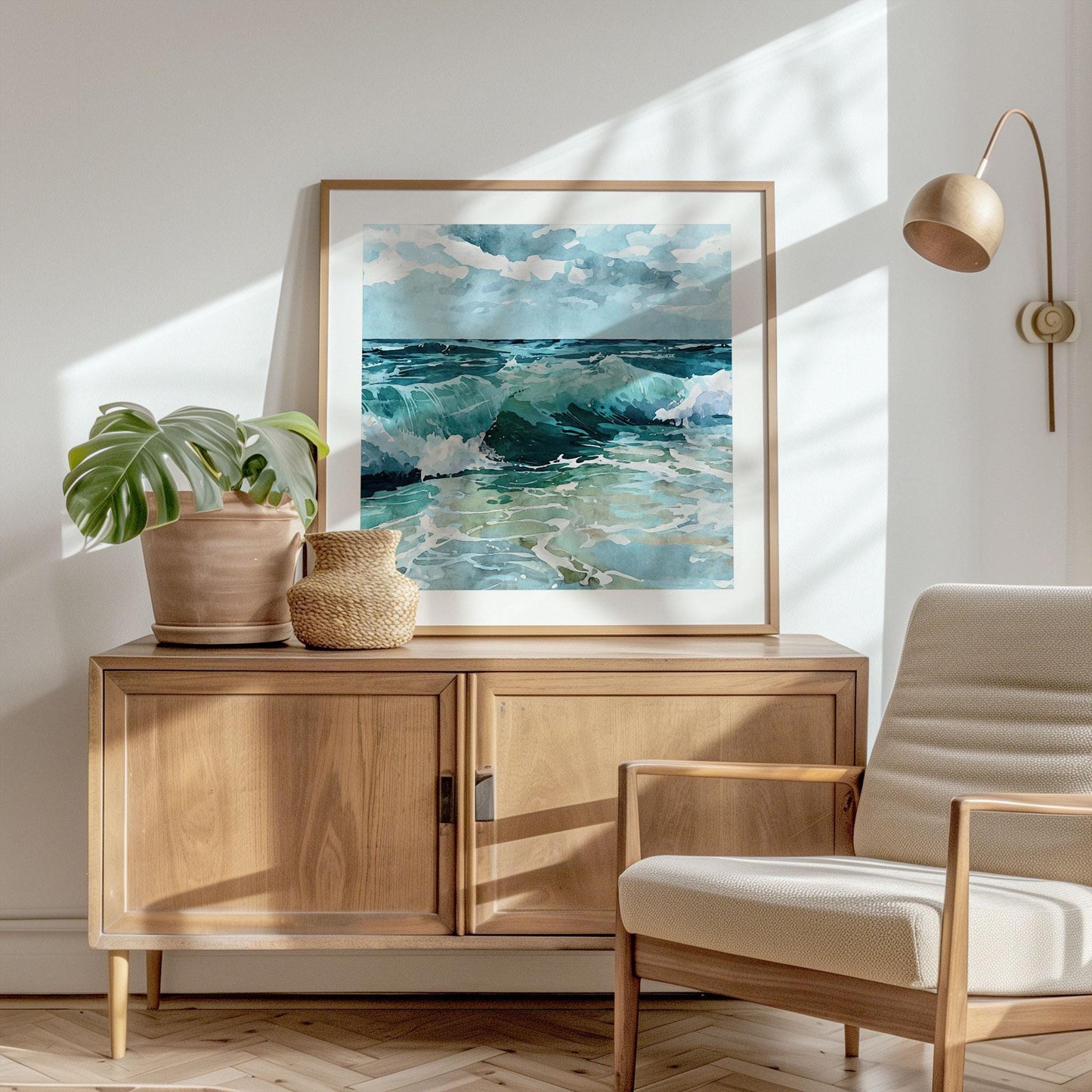 Framed watercolor painting of ocean waves in shades of turquoise, teal, and blue rests on a wooden sideboard, styled with a potted plant and woven basket in a sunlit modern living room with a neutral armchair and wall-mounted lamp.