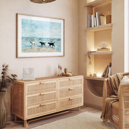 Framed watercolor-style painting of three Labrador dogs, one yellow and two black, walking along a sandy beach with blue sky and waves, hung above a light wood rattan dresser in a cozy, neutral-toned living space with warm lighting, wicker furniture, and decorative accents.