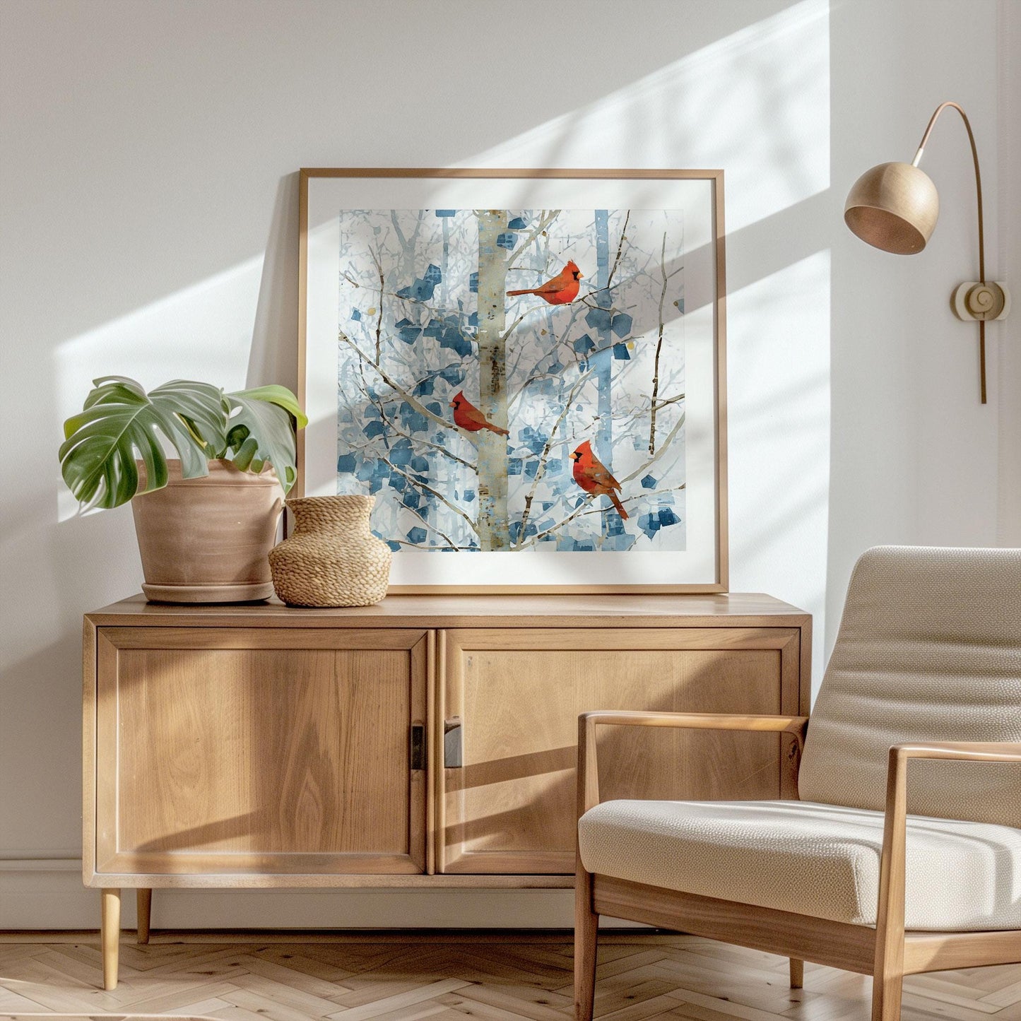 Framed square art print of three red cardinals perched on pale birch branches with soft blue winter foliage, displayed on a light wood sideboard. A potted monstera and woven vase sit beside it, with a mid-century style armchair and warm sunlight casting gentle shadows across the bright, minimalist room.