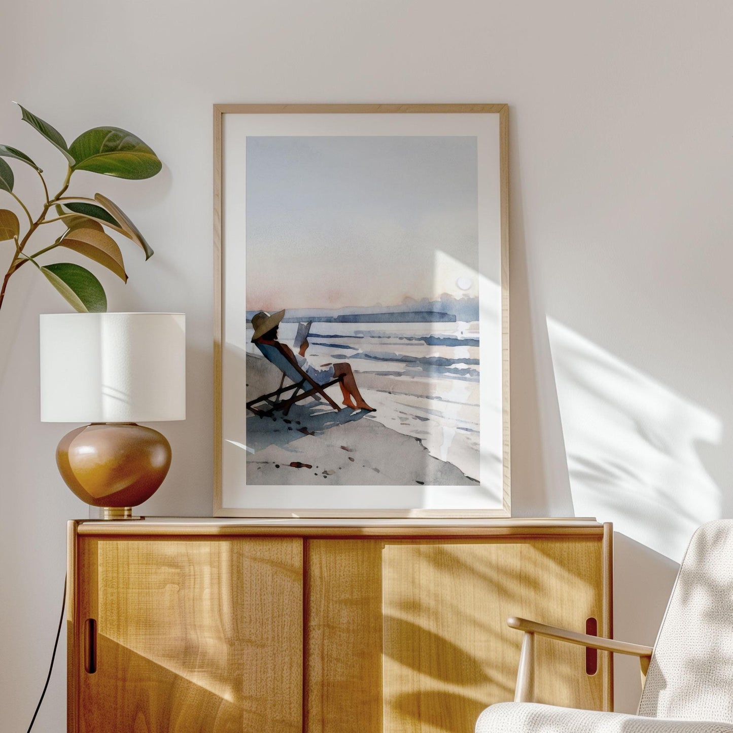 Framed watercolor painting of a woman in a sun hat reading by the ocean, displayed on a light wood cabinet. A ceramic lamp and potted plant sit nearby, with soft sunlight casting natural shadows across the wall and furniture, creating a calm, coastal-inspired interior.