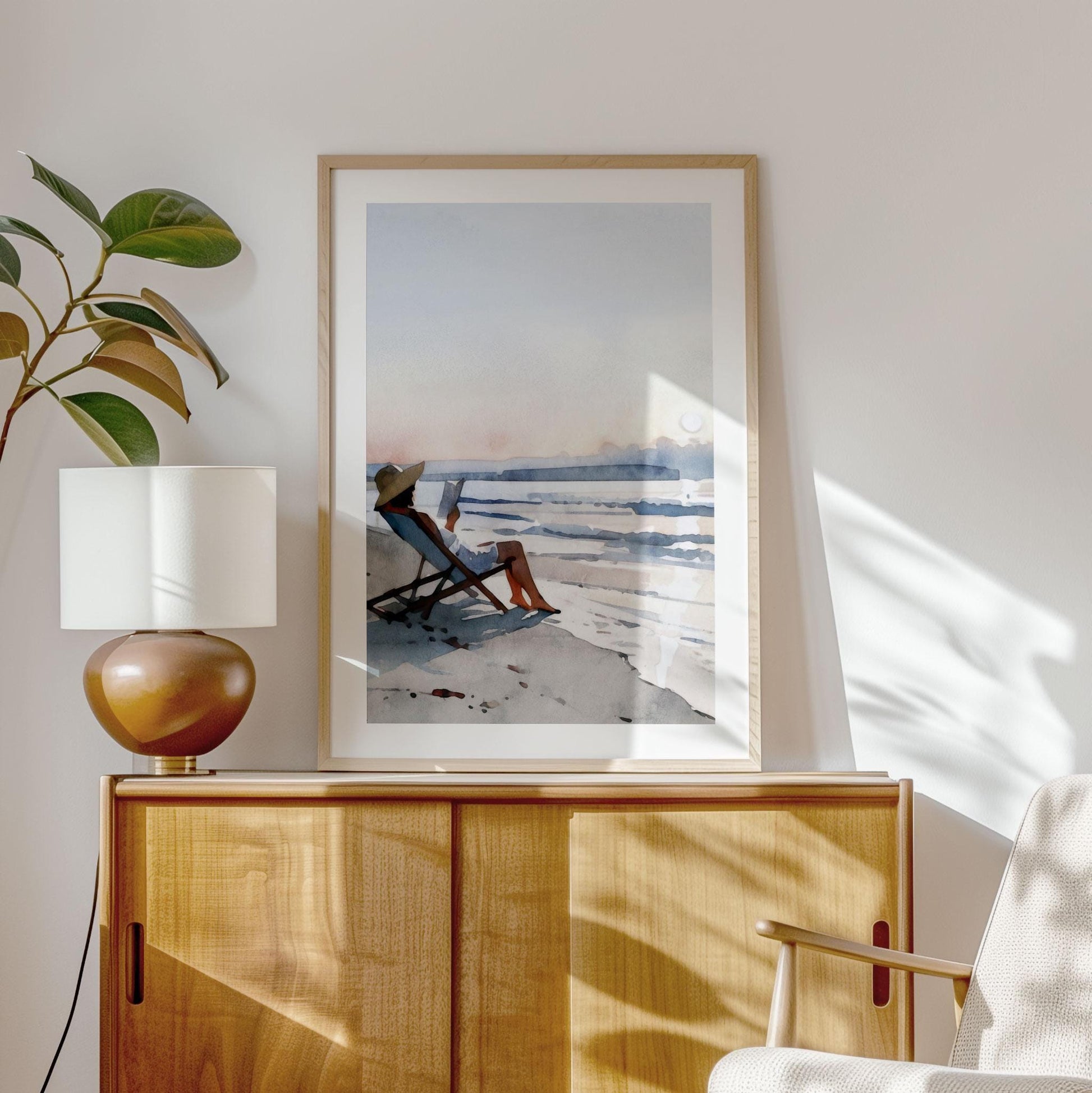 Framed watercolor painting of a woman in a sun hat reading by the ocean, displayed on a light wood cabinet. A ceramic lamp and potted plant sit nearby, with soft sunlight casting natural shadows across the wall and furniture, creating a calm, coastal-inspired interior.