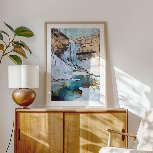 Framed watercolor painting of a snowy waterfall scene with turquoise pool, displayed on a light wood cabinet beside a modern lamp and plant, in a sunlit minimalist living room.