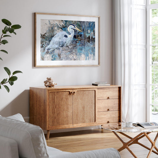 Framed watercolor-style heron print hangs above a mid-century wooden sideboard with woven doors, in a bright living room with white walls, light wood floors, and sheer curtains letting in natural light.