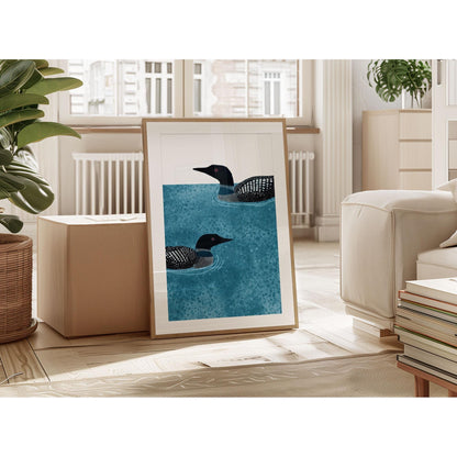 Framed illustration of two loons swimming in mottled blue water, leaning against a wall in a bright living room. The artwork rests on the floor near a beige sofa, stacked books, and a cardboard box, with large windows letting in natural light and green plants adding a cozy touch.