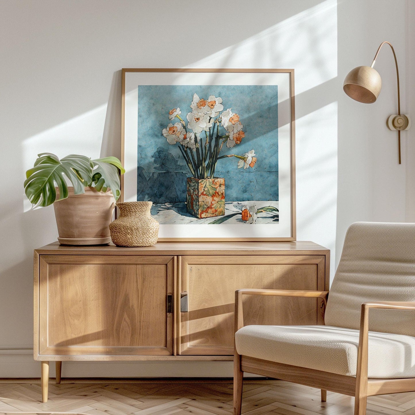 Framed floral art print of white daffodils with orange centers in a patterned vase against a textured blue background, styled on a wooden sideboard with a potted monstera, woven basket, and neutral armchair, in a sunlit modern interior.
