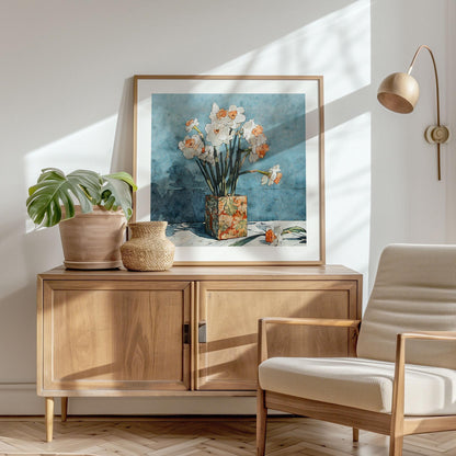 Framed floral art print of white daffodils with orange centers in a patterned vase against a textured blue background, styled on a wooden sideboard with a potted monstera, woven basket, and neutral armchair, in a sunlit modern interior.