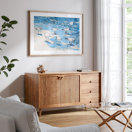 Framed coastal watercolor art print featuring a group of small white shorebirds with orange beaks and legs, standing and wading in shallow blue water along a sandy shoreline. The fluid waves and soft sandy patches are outlined in black ink for a lively, illustrative style. Displayed above a wooden cabinet with woven rattan doors in a bright, airy living room with natural light streaming through sheer white curtains.