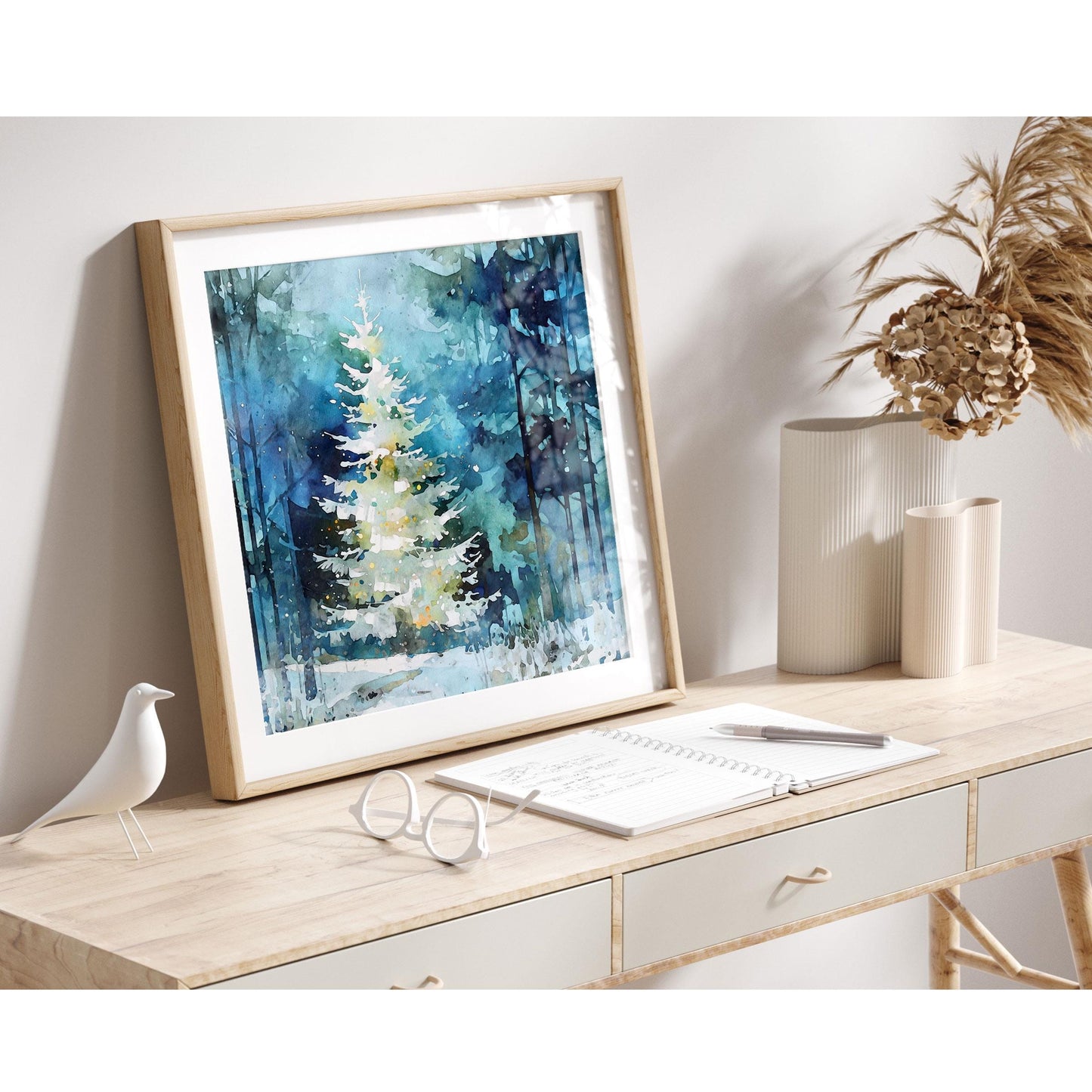 A framed watercolor print of a glowing white Christmas tree in a snowy blue-green forest rests on a light wooden desk. Neutral décor surrounds it—ribbed cream vases, dried foliage, a minimalist white bird figurine, and an open notebook with glasses—creating a calm, modern, Scandinavian-inspired holiday scene.