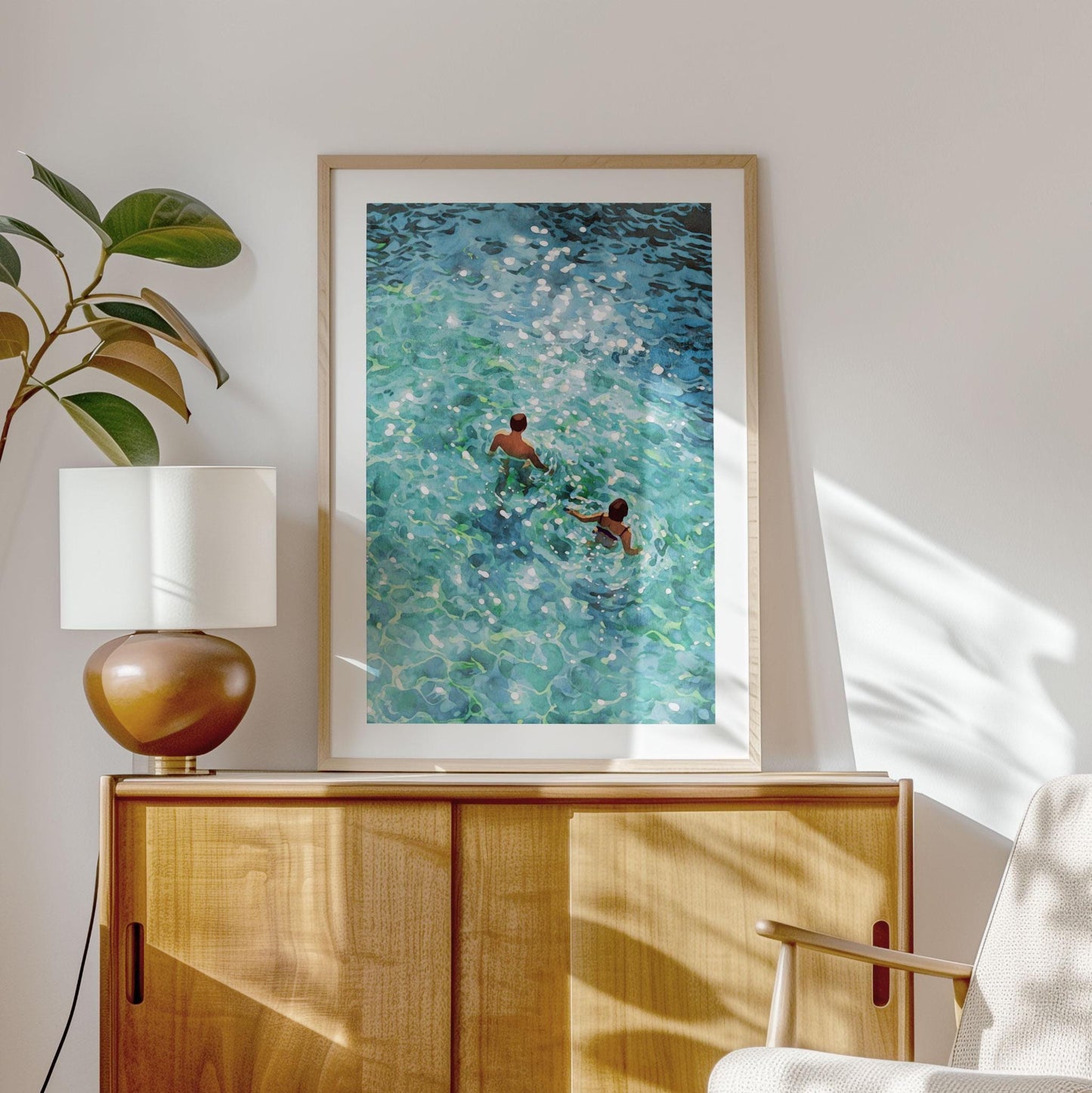 Framed painting of two swimmers viewed from above, moving through clear turquoise water with sparkling sunlight reflections, displayed on a light wooden cabinet beside a modern lamp and plant in a bright, sunlit room.