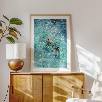 Framed painting of two swimmers viewed from above, moving through clear turquoise water with sparkling sunlight reflections, displayed on a light wooden cabinet beside a modern lamp and plant in a bright, sunlit room.