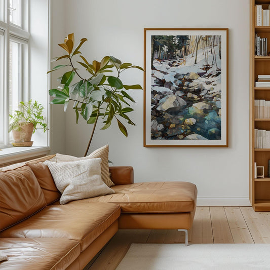 Modern living room with a tan leather sectional sofa and neutral throw pillows, a tall potted plant near a large window, and a framed watercolor-style painting of a snowy forest stream with rocks and reflections hanging on the wall. A wooden bookshelf with neatly arranged books completes the cozy, minimalist setting.
