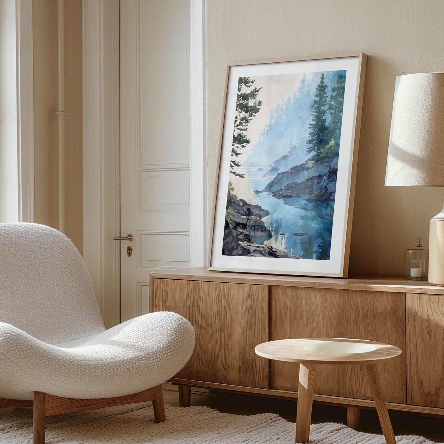 Styled interior photo showing a framed watercolor-style landscape art print leaning on a wooden sideboard in a calm, neutral living space. The artwork depicts a misty blue lake with rocky shoreline and evergreen trees reflected in still water. Soft natural light, light wood furniture, and a textured armchair create a warm, modern, minimalist home decor setting.