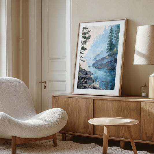 Styled interior photo showing a framed watercolor-style landscape art print leaning on a wooden sideboard in a calm, neutral living space. The artwork depicts a misty blue lake with rocky shoreline and evergreen trees reflected in still water. Soft natural light, light wood furniture, and a textured armchair create a warm, modern, minimalist home decor setting.