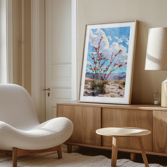 Framed desert landscape painting of a flowering ocotillo with red blooms set against a bright blue sky and distant purple mountains, displayed on a wooden sideboard in a modern beige living room. The space features a curved white textured chair, wooden table, and soft natural lighting creating a warm, minimalist aesthetic.