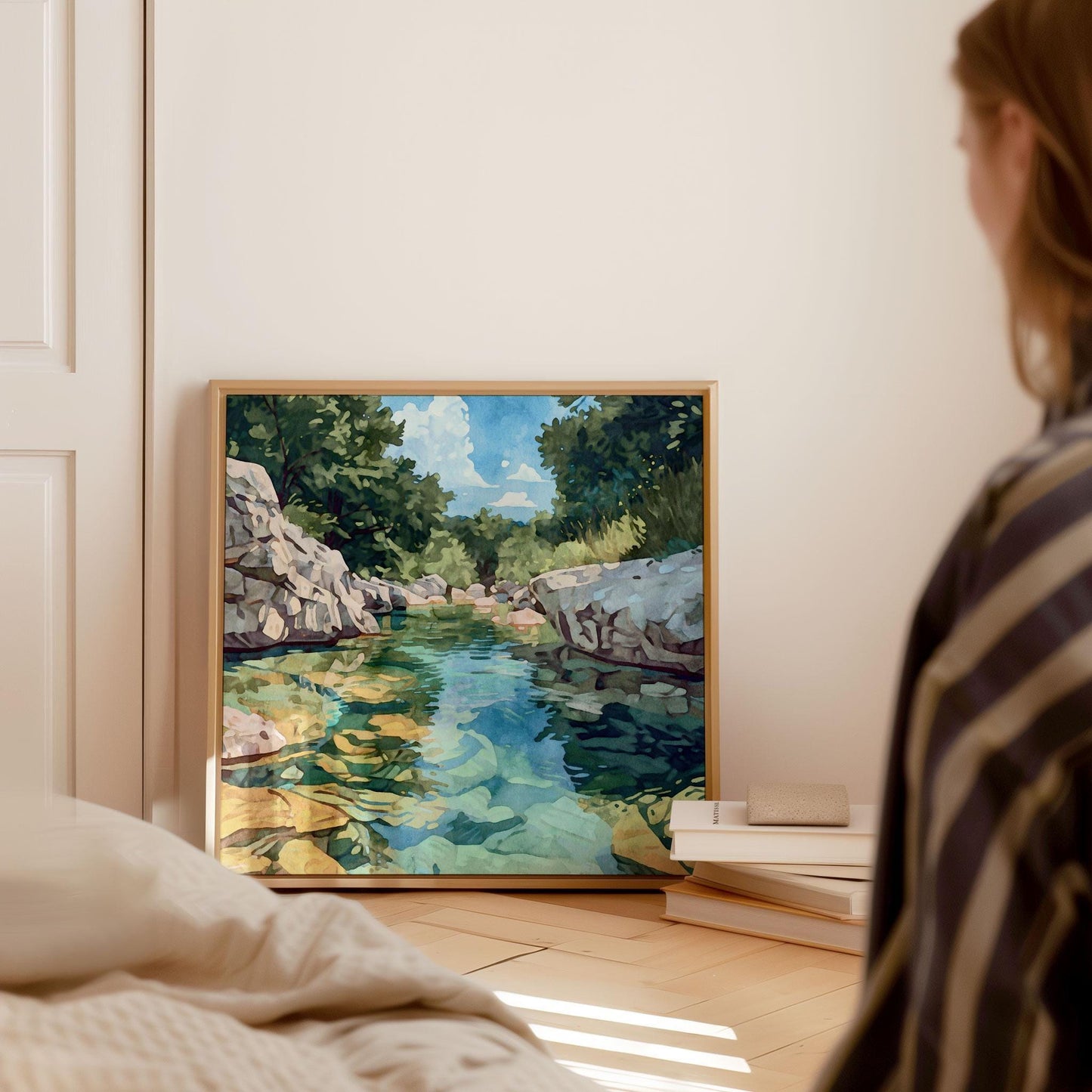 Framed watercolor-style painting of a clear rocky river with turquoise and green water, leaning against a wall beside stacked books in a softly lit bedroom. A woman in striped clothing sits nearby, gazing at the artwork, with sunlight streaming across the wooden floor.
