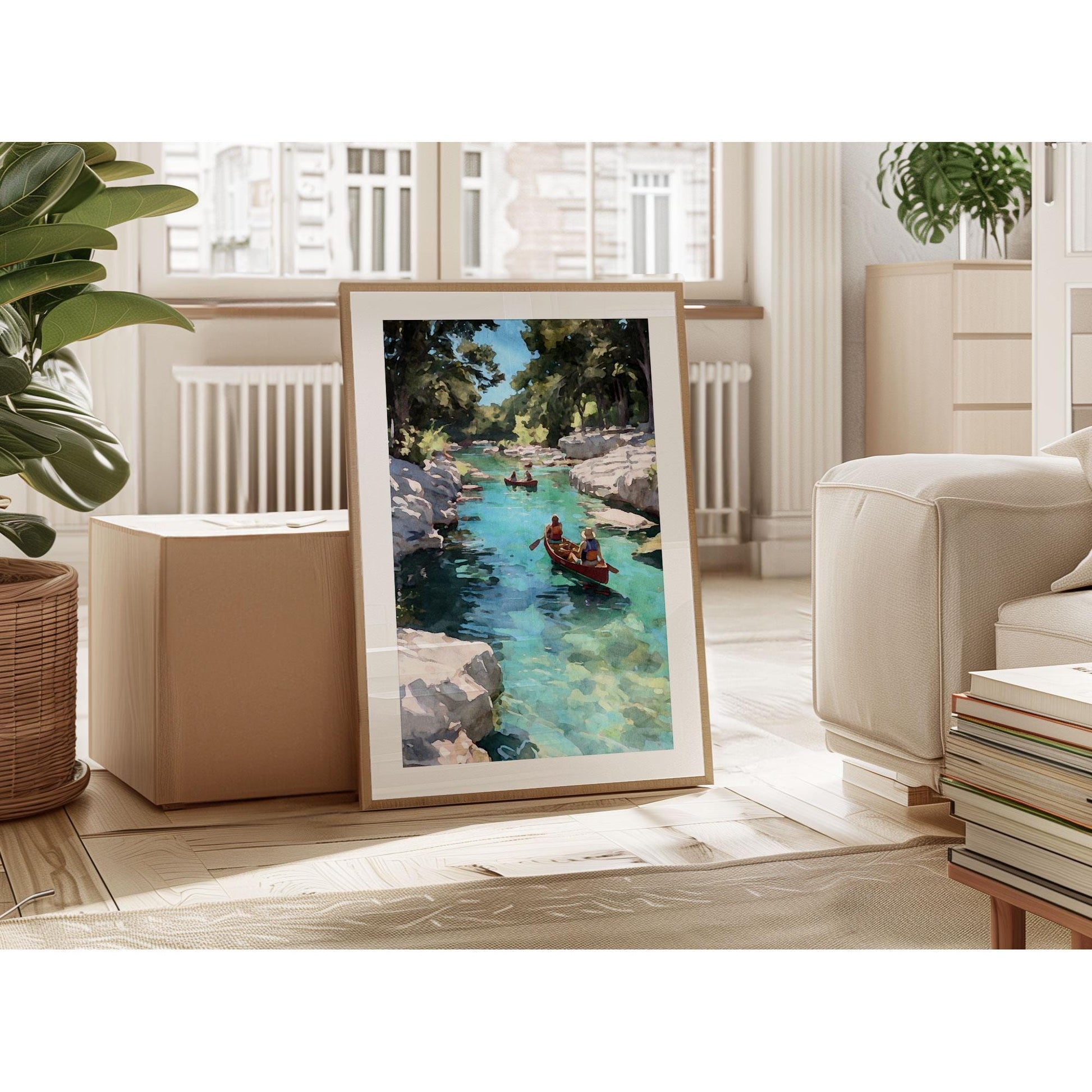 Framed painting of two people canoeing on a turquoise river surrounded by rocky cliffs and trees, displayed leaning against a wall in a bright, modern living room with plants, books, and natural light from large windows.