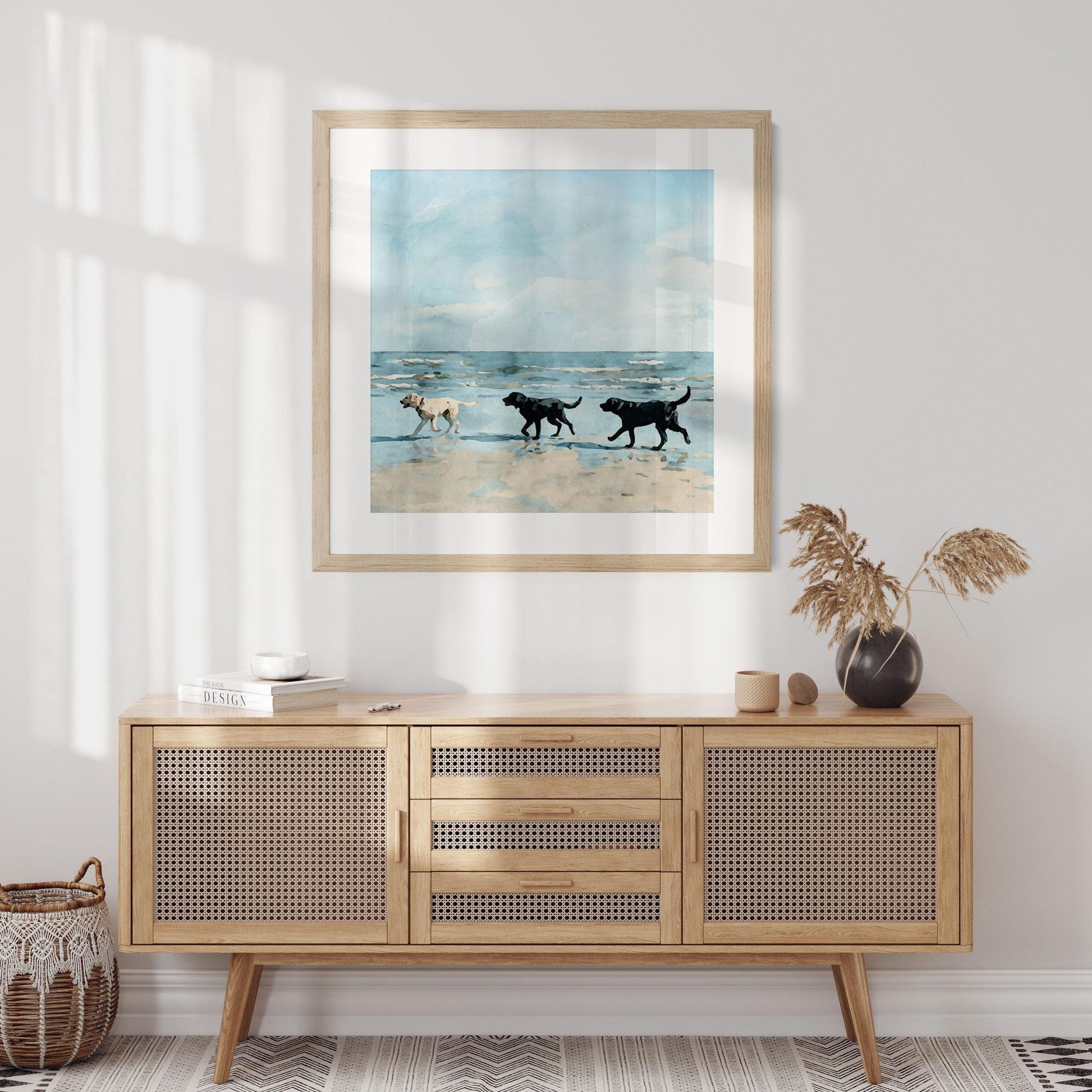 Framed watercolor-style painting of three Labrador dogs, one yellow and two black, walking along a sandy beach with gentle waves and a soft blue sky, displayed on a white wall above a mid-century modern rattan-front sideboard with neutral decor accents and dried grasses in a black vase.