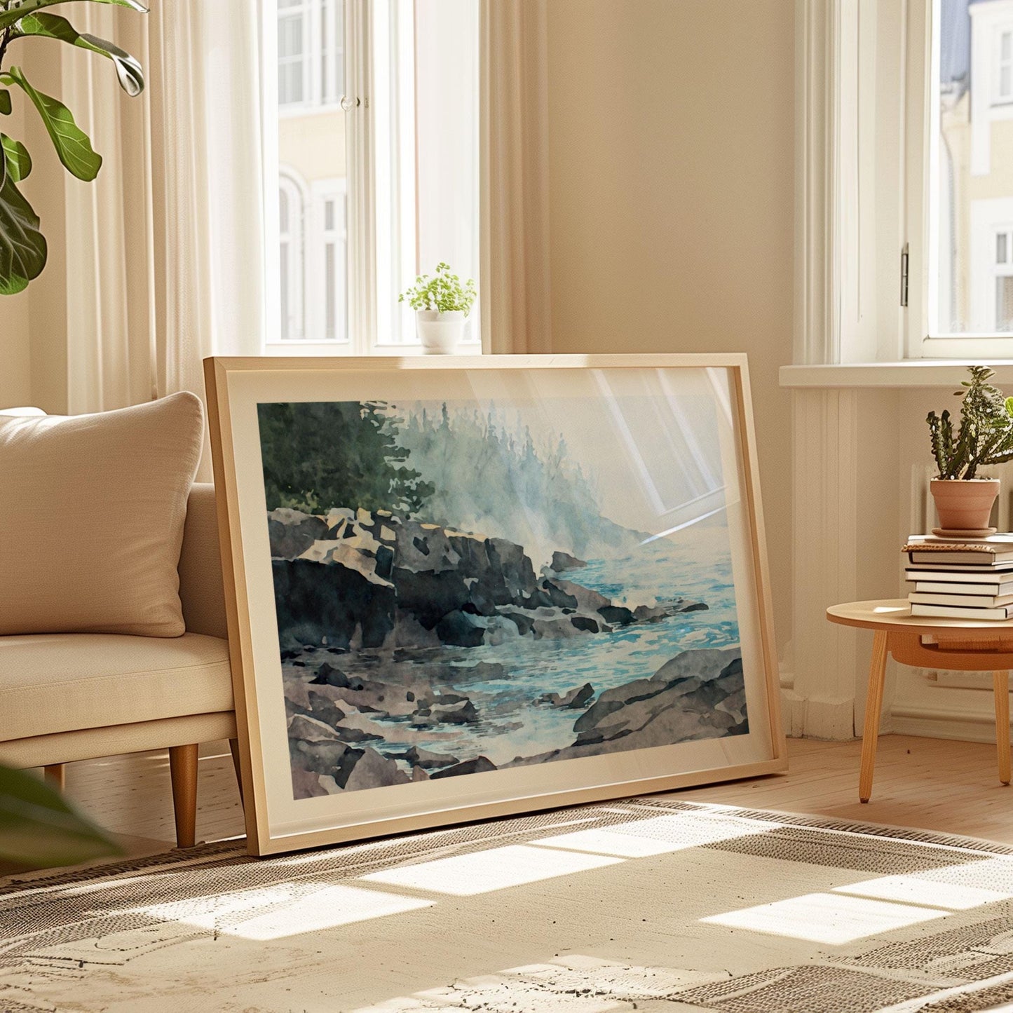 Framed horizontal art print leaning against a wall in a sunlit living room. The watercolor-style artwork depicts a misty Lake Superior shoreline with dark rocky cliffs, calm blue water, and soft fog drifting across a distant evergreen forest. Warm natural light, neutral furnishings, and indoor plants create a serene, cozy interior setting that highlights the artwork’s muted blues, grays, and greens.