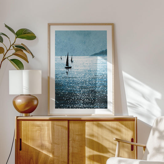 Framed sailboat art print displayed on a light wood cabinet in a sunlit room. The artwork features dark sailboats gliding across shimmering blue water, with sparkling highlights and a misty shoreline in the distance. A matte border surrounds the print. The modern, coastal decor includes a round ceramic lamp, leafy plant, and a mid-century style armchair, creating a calm, inviting atmosphere.