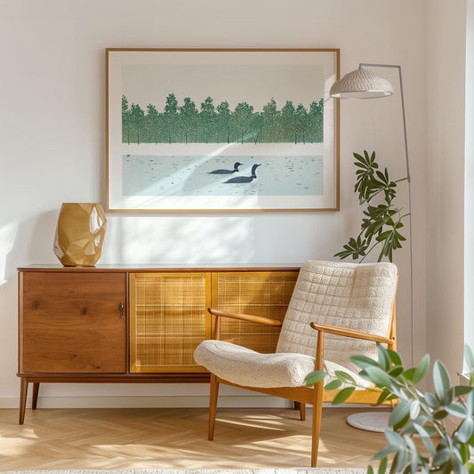 Framed loon art print hanging above a mid-century wood sideboard in a sunlit living room. The illustration features two loons gliding on a calm lake with a line of stylized green trees in the background. Cozy modern decor includes a textured lounge chair, sculptural gold vase, and leafy houseplants.