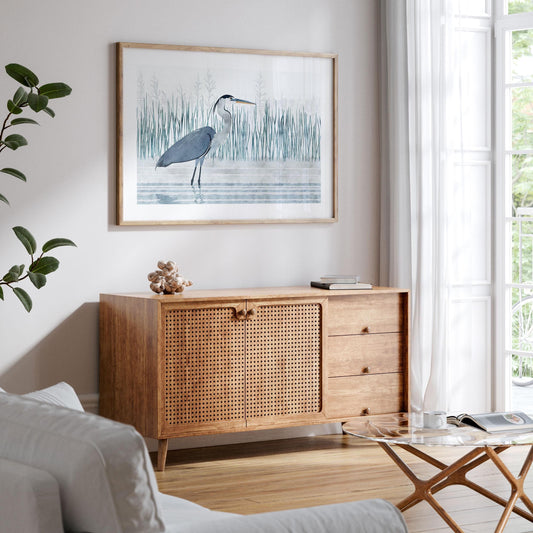 Framed watercolor-style print of a great blue heron standing in water, hung above a mid-century modern wood cabinet with rattan doors. The art features tall green reeds and soft gray tones. The scene is set in a bright, minimalist living room with white walls, sheer curtains, and natural light pouring through a large window. A light gray sofa, glass coffee table, and potted plant complete the airy, coastal-inspired decor.