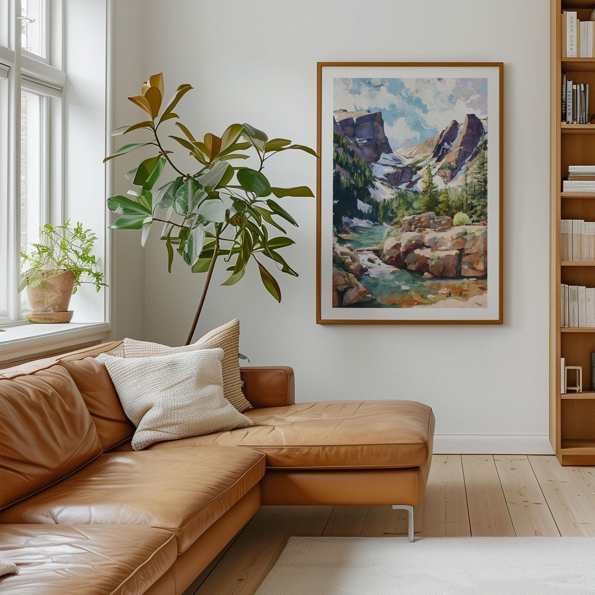 cccFramed mountain landscape painting displayed in a modern living room with a tan leather sectional sofa, cream pillows, and a large leafy plant by a bright window. The artwork shows rugged peaks, pine forests, and a turquoise river under a cloudy sky, painted in a modern impressionist style. The natural wood frame complements the room’s light wood floors, soft neutral tones, and minimalist decor.