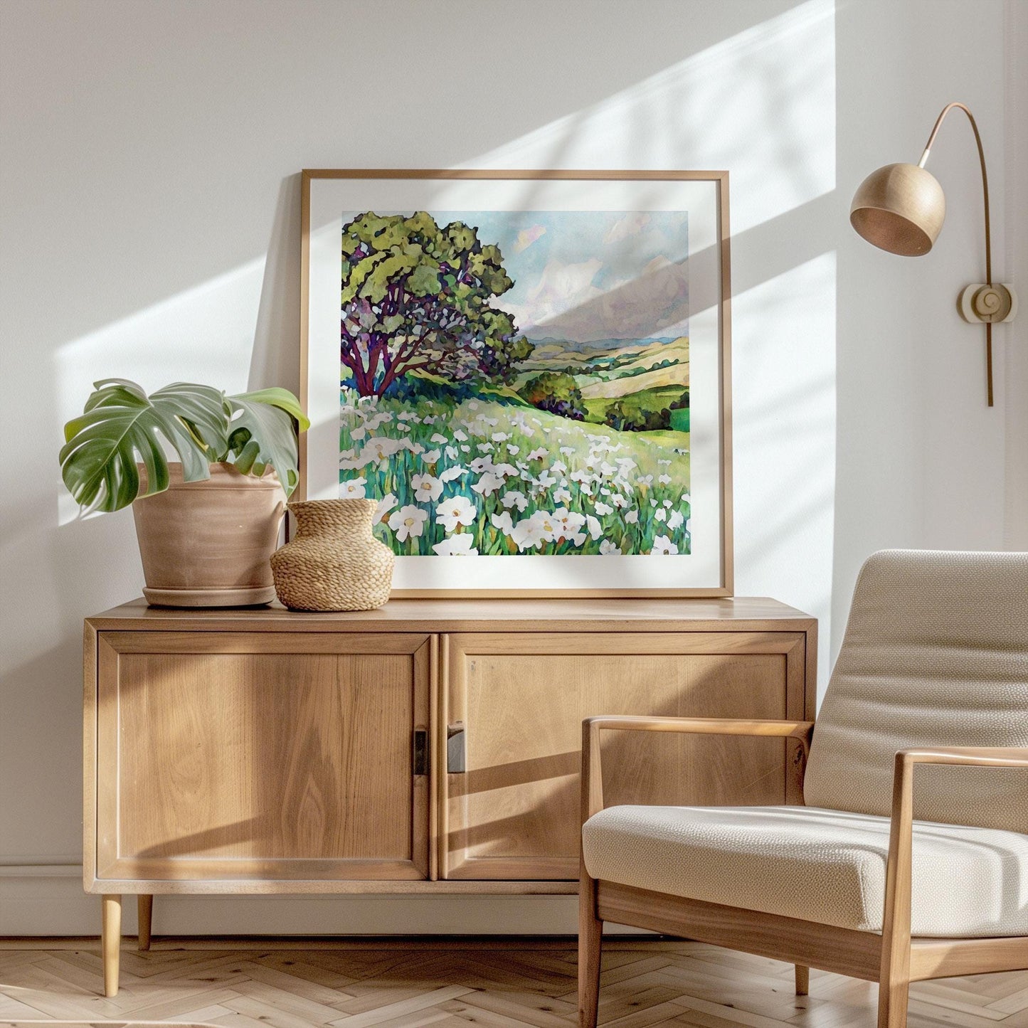 Framed square painting of a sunny meadow with white wildflowers and a large tree, overlooking rolling green hills under a blue sky, displayed on a wooden sideboard beside potted plants in a bright, modern room with natural light streaming in.