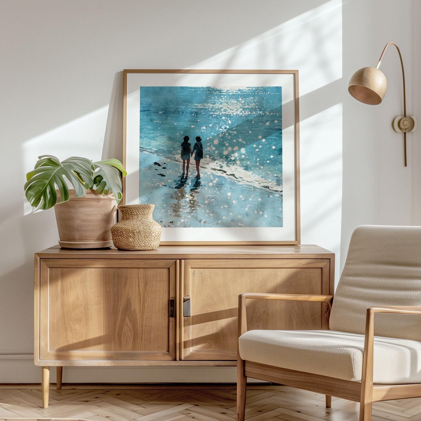 Framed beach watercolor print styled in a sunlit, modern interior with light wood furniture and neutral decor. The artwork shows two women holding hands at the water’s edge, surrounded by glistening sea spray and soft blue waves.