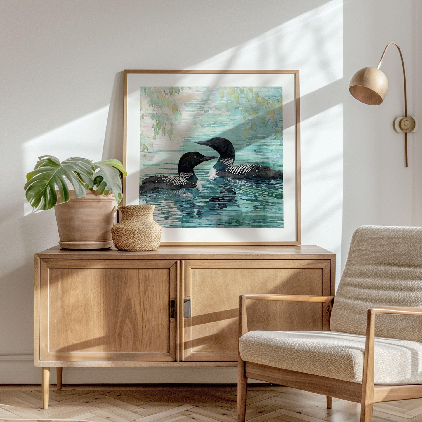 Framed watercolor painting of two loons gliding on a lake, displayed on a wooden cabinet in a sunlit room. The artwork shows black-and-white loons with soft reflections on blue-green water, accented by leafy branches above. A potted monstera plant, woven basket, and modern beige armchair complete the warm, minimalist decor.